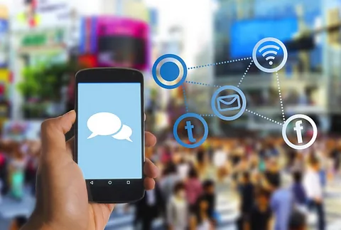 Eine Hand hält ein Smartphone mit einer Sprechblase auf dem Bildschirm, umgeben von Social-Media-Symbolen vor einem verschwommenen Stadtbild