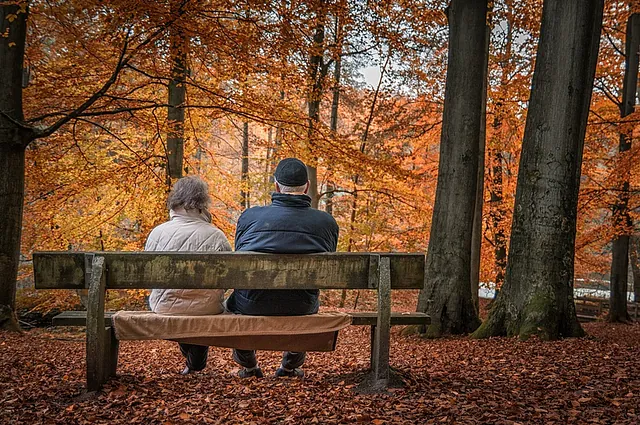 Ein älteres Paar sitzt auf einer Bank in einem herbstlichen Wald und blickt nach vorn.