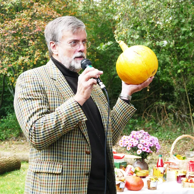 Ein älterer Mann mit Bart hält eine gelbe Kugel hoch und spricht in ein Mikrofon im Freien